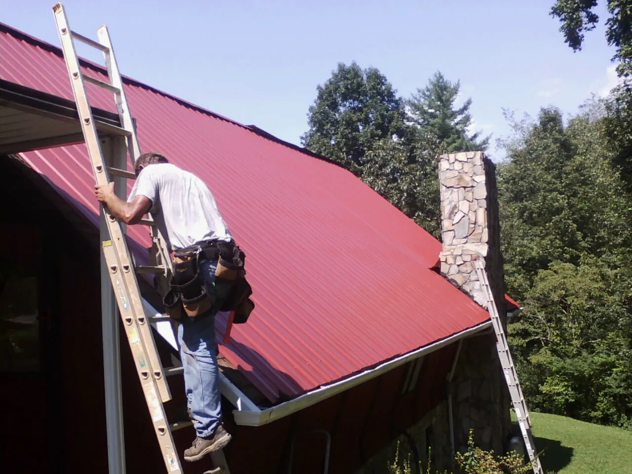 Roofer on a ladder beside a red metal roof and stone chimney — Soffit & Fascia Repair by Roofing Brothers in Roachdale